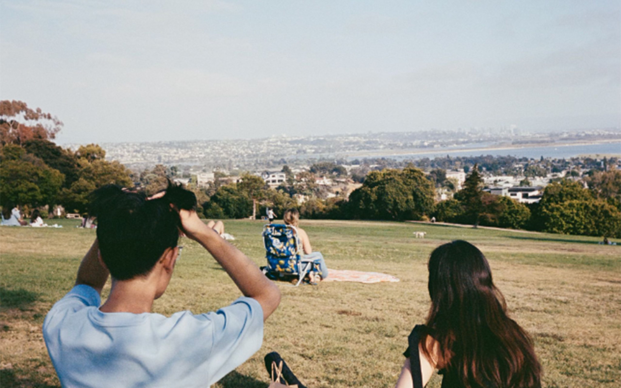 Two figures, one male and one female sitting on top of a hill that overlooks a coastline and mountains. There is a person in a lawn chair sitting in front of them.